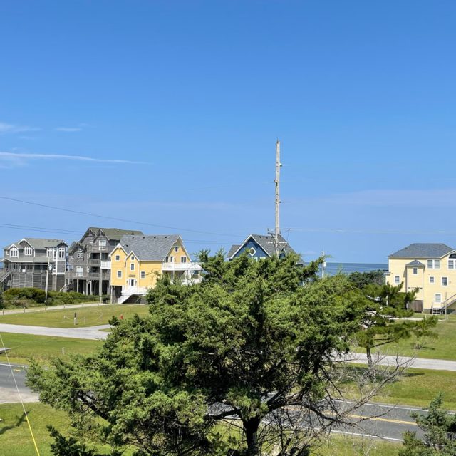 Several colorful houses and trees line a street under a clear blue sky near the ocean.