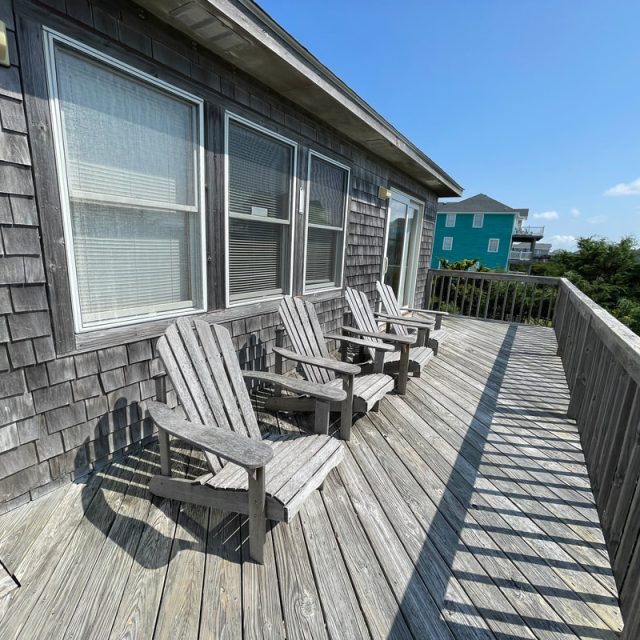 Four wooden chairs sit on a sunlit wooden deck attached to a house, with neighboring houses in the background.