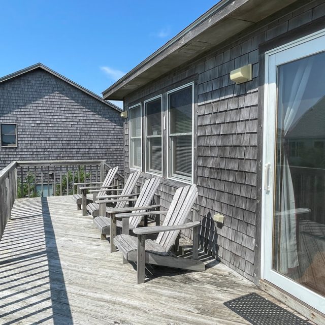 Four wooden chairs on a sunlit deck beside a house with shingled siding, under a clear blue sky.