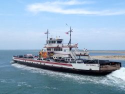 Car ferry named Hatteras crossing water with vehicles on board, against a backdrop of sky and sandy shore.