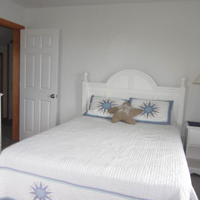 A white bed with star-themed pillows, a nightstand with a lamp, and a dresser with flowers in a bright bedroom.