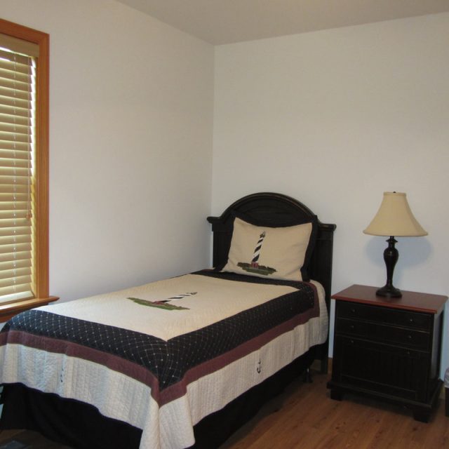 Small bedroom with a single bed, lighthouse-themed bedding, nightstand, lamp, window with blinds, and trash bin.