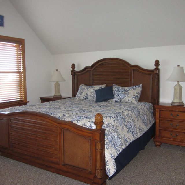 Bedroom with a large wooden bed, matching nightstands, two lamps, and windows with blinds on the left.