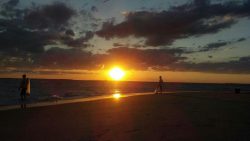 Two people walk on a beach at sunset, with the sun low over the horizon and clouds in the sky.