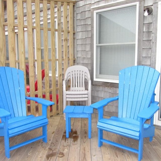 Two blue wooden chairs and a small table on a wooden deck; a white plastic chair is behind them near the house.