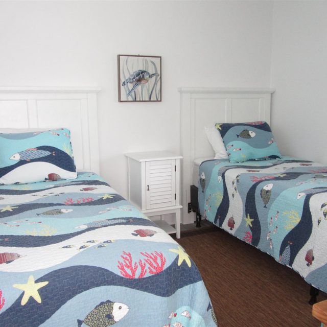 Two twin beds with fish-patterned bedding and a small white nightstand between them in a simple, white room.