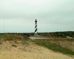 A black and white striped lighthouse stands near a sandy area with grass and a cloudy sky overhead.