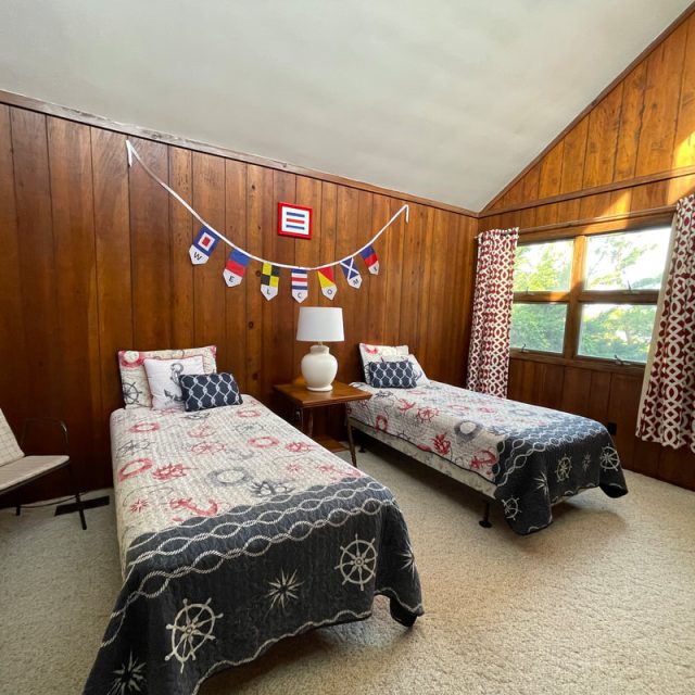 Bedroom with two twin beds, nautical bedding, wood-paneled walls, and a window with patterned curtains.
