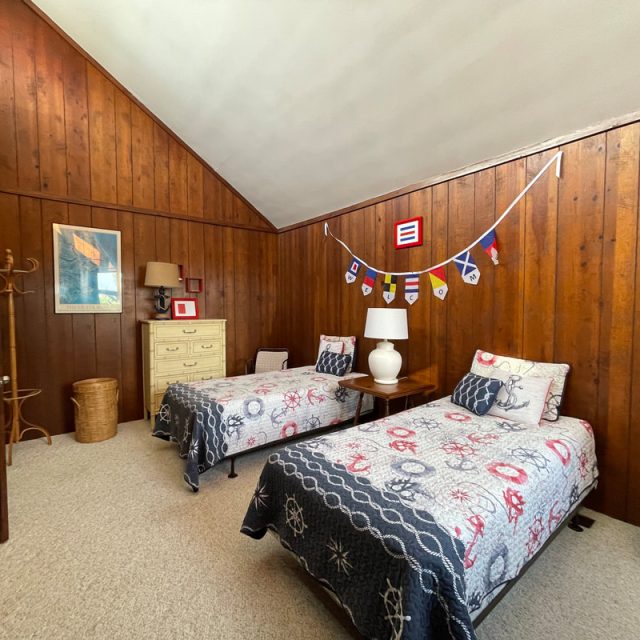 Wood-paneled bedroom with two twin beds, nautical decor, dresser, lamp, and coat rack on beige carpeted floor.