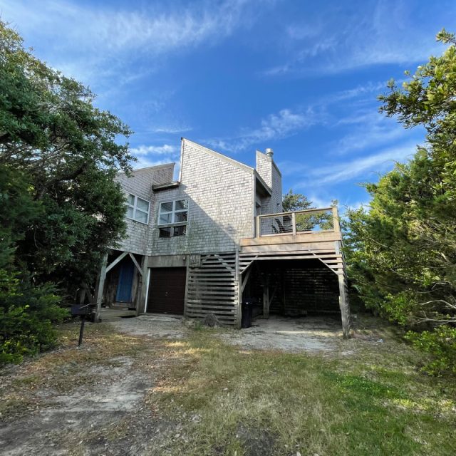 Two-story wooden house with a deck, surrounded by trees, under a blue sky with scattered clouds.