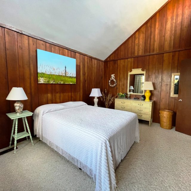 Bedroom with wood-paneled walls, a bed with a white cover, lamps, a dresser with a mirror, and a wicker hamper.