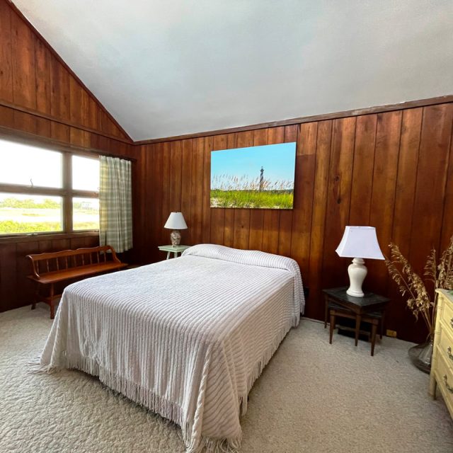 Wood-paneled bedroom with a bed, two lamps, a dresser, a bench by the window, and a landscape painting above the bed.