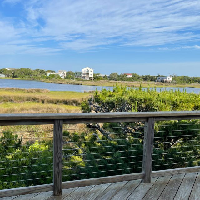 Wooden deck with railing overlooks grassy marsh, water, and distant houses under a blue sky with scattered clouds.