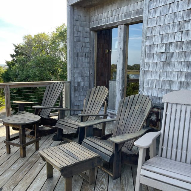 Four wooden Adirondack chairs and a small table sit on a weathered deck outside a shingled house, surrounded by greenery.