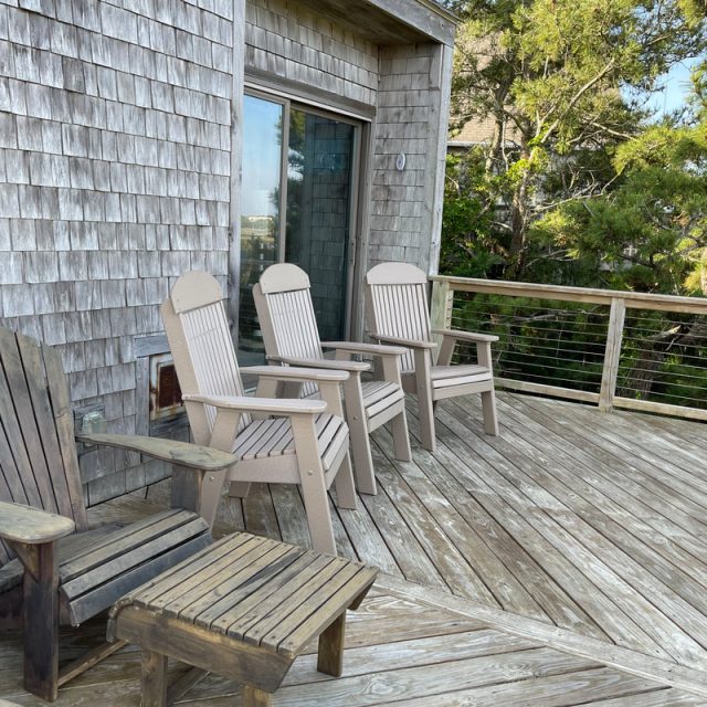 Four wooden chairs and two footstools on a weathered deck outside a shingled house, surrounded by trees.
