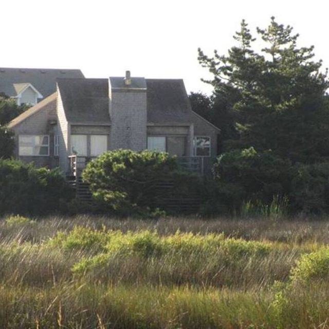 A small, gray, weathered house sits among trees and tall grass with other houses nearby in the background.