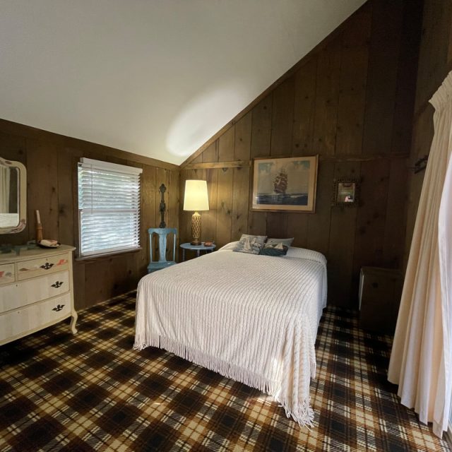Bedroom with wood-paneled walls, plaid carpet, a bed, dresser, lamp, and window with curtains.