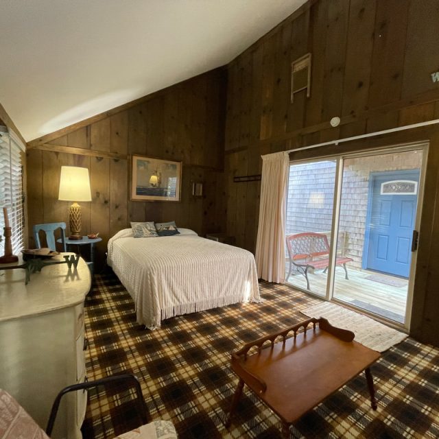 Wood-paneled bedroom with plaid carpet, bed, dresser, armchair, and sliding glass door to outdoor seating area.