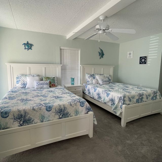 Bedroom with two white beds, sea-themed bedding, a ceiling fan, and ocean decor on pale walls.