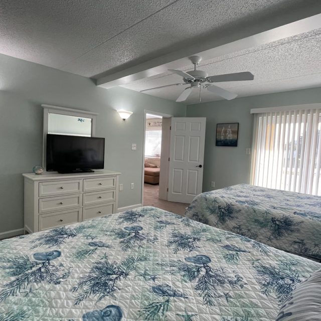 Bedroom with two beds, TV on dresser, ceiling fan, and sliding glass doors with vertical blinds letting in sunlight.