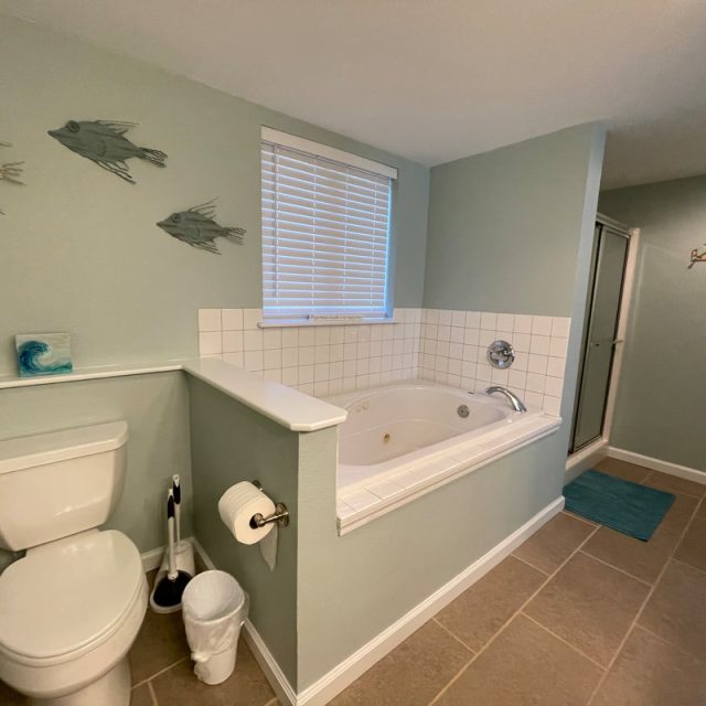 Bathroom with toilet, whirlpool tub, wall art of fish, tiled floor, shower, window with blinds, and wall hooks.