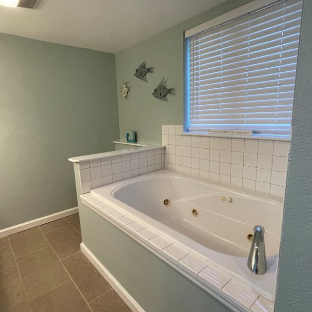 Large white bathtub with jets, tiled surround, fish wall decor, and a window with blinds in a bathroom.