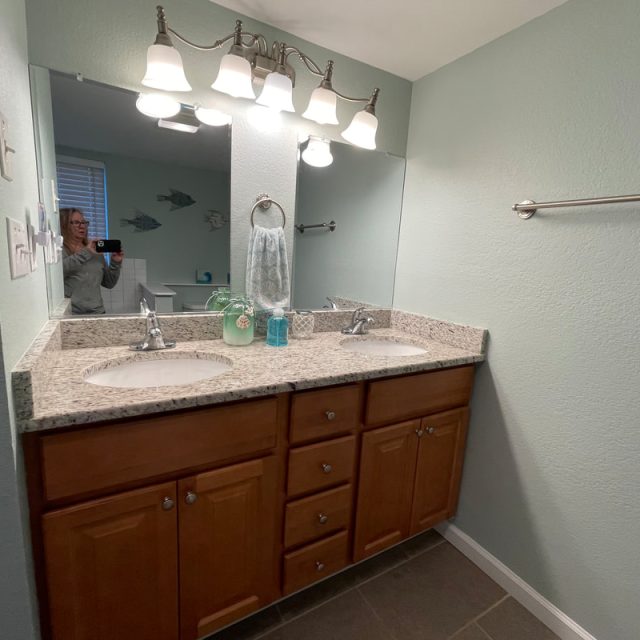 Modern bathroom with double sink vanity, granite countertop, wall mirror, and light fixture; person visible in mirror.