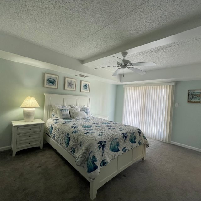 Bedroom with a white bed, nightstand with lamp, ceiling fan, carpet, and sliding door with vertical blinds.