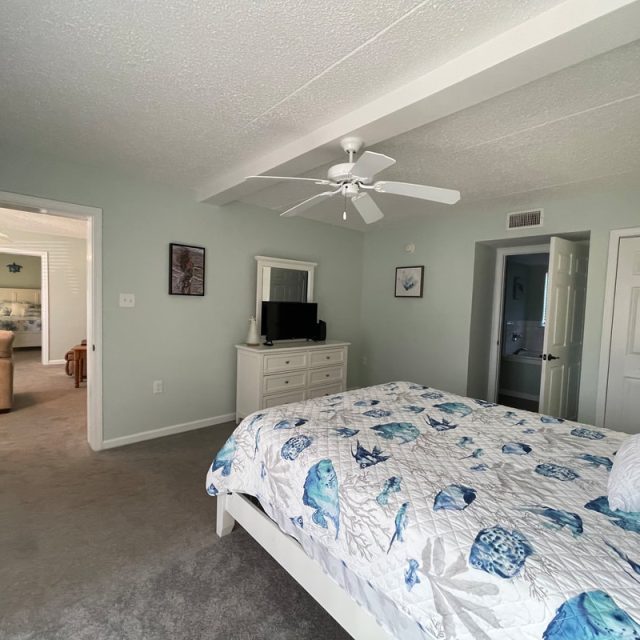 Bedroom with a seashell-patterned bedspread, dresser with TV, ceiling fan, and open doorways to other rooms.