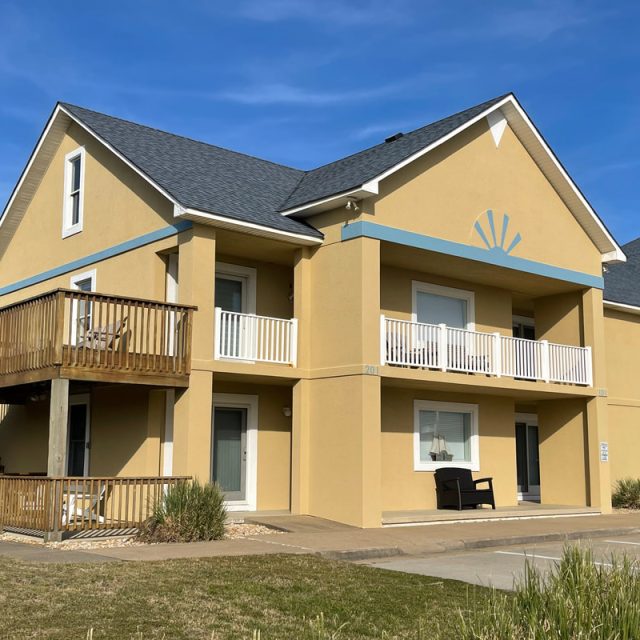 Two-story yellow building with white balconies, wooden deck, and outdoor seating under a clear blue sky.