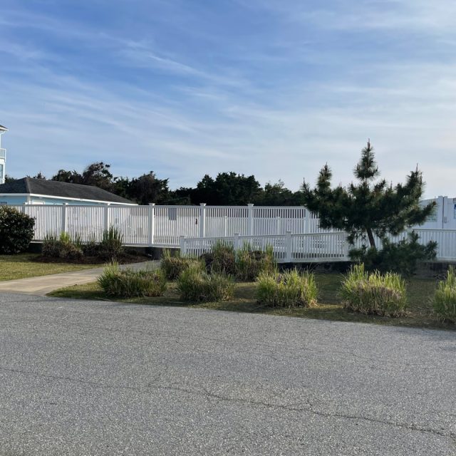 White fence surrounds a pool area next to a house, with shrubs and trees along a quiet paved street under a blue sky.
