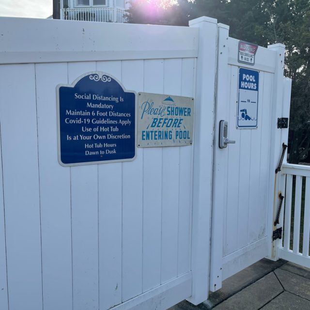 White pool gate with signs about social distancing, showering before entering, and posted pool hours from dawn to dusk.