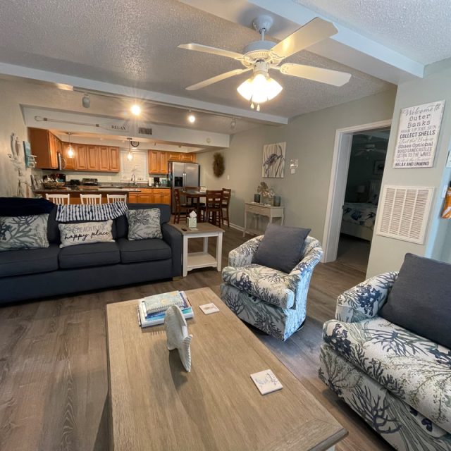 Living room with sofa, two armchairs, ceiling fan, coffee table, and view of kitchen in the background.