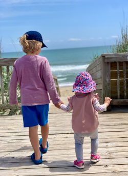 Two young children walk hand in hand on a wooden boardwalk toward the beach on a sunny day.