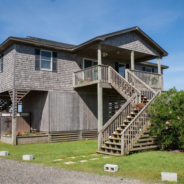 Two-story wooden house on stilts with an exterior staircase, set on a grassy lawn under a clear blue sky.