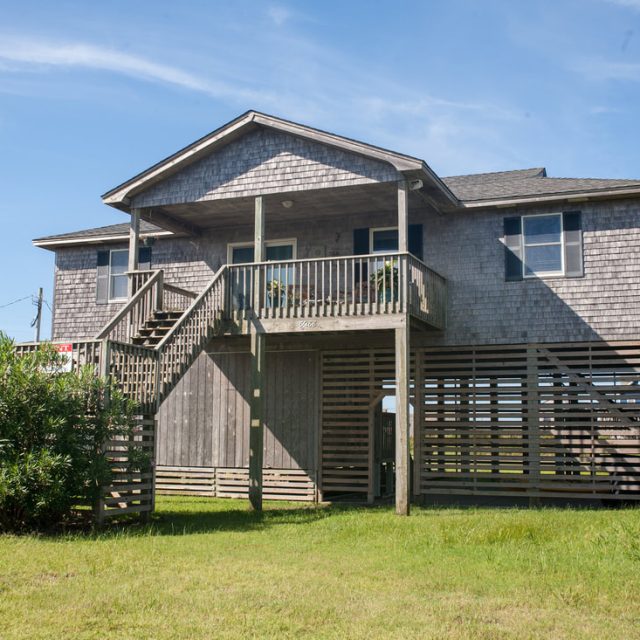A gray wooden beach house on stilts with a front porch, stairs, and grassy yard under a clear blue sky.