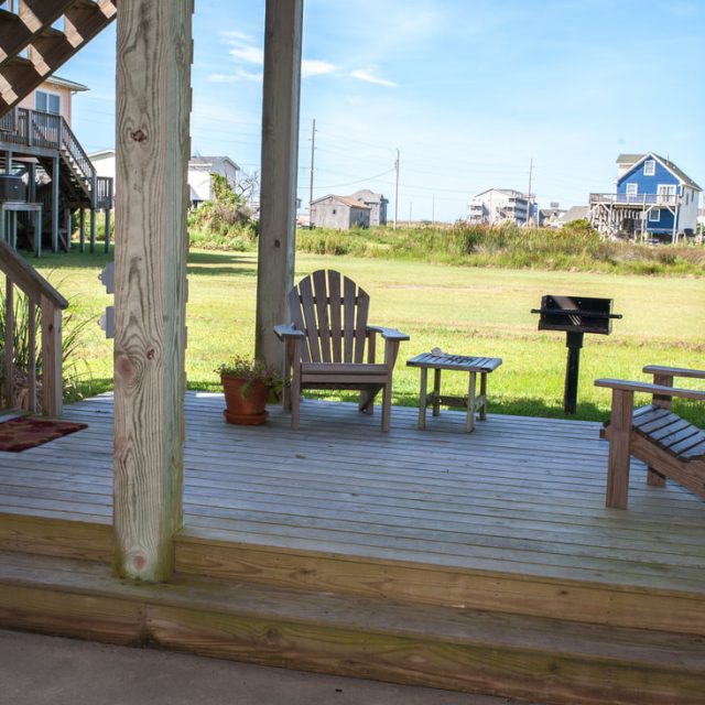 Wooden patio with two chairs, a small table, potted plants, and a grill, overlooking a grassy area and houses.