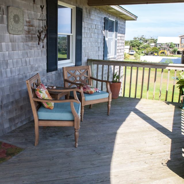 Two wooden chairs with cushions sit on a sunny wooden porch with potted plants and a view of neighboring houses.