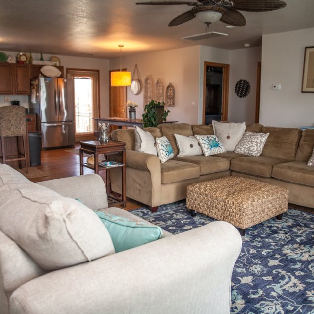 Open living room with two beige sofas, a wicker coffee table, blue rug, and a kitchen in the background.