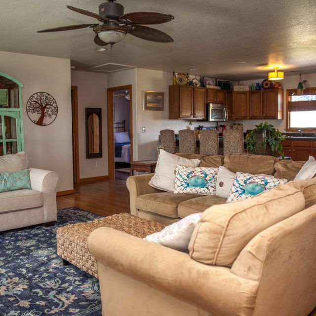 Living room with tan couches, blue rug, and open kitchen in the background with wood cabinets and stainless steel fridge.