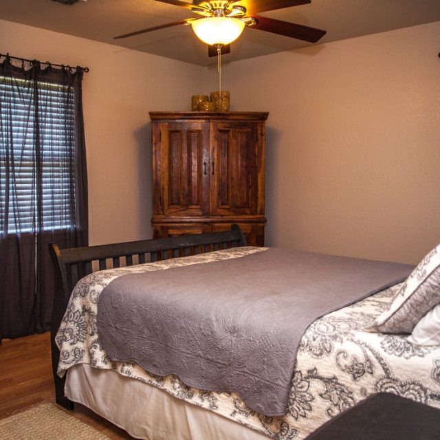 Bedroom with a wooden bed, patterned bedding, wooden armoire, two windows with dark curtains, and a ceiling fan.