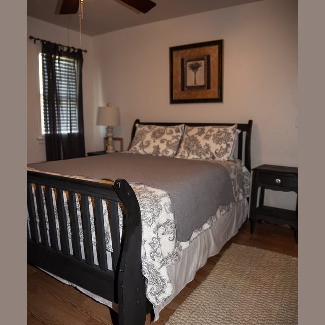 Bedroom with a dark wood bed, patterned bedding, nightstand, lamp, and framed art on a white wall.