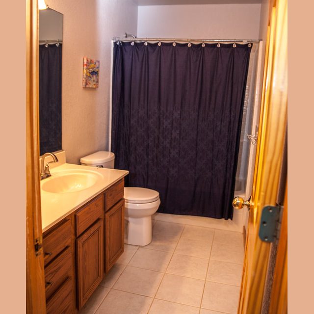 Bathroom with wooden vanity, white sink, toilet, tiled floor, and dark shower curtain behind the toilet.