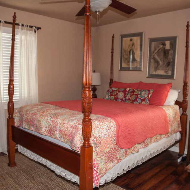 A bedroom with a four-poster bed, coral bedding, two windows with white curtains, and framed art on the wall.