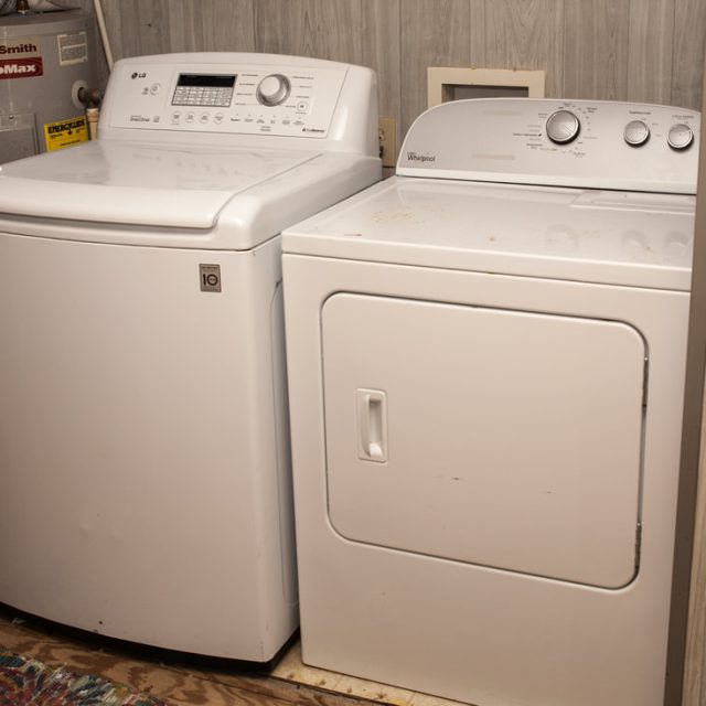A white top-load washing machine and a front-load dryer side by side in a small laundry area with wood panel walls.