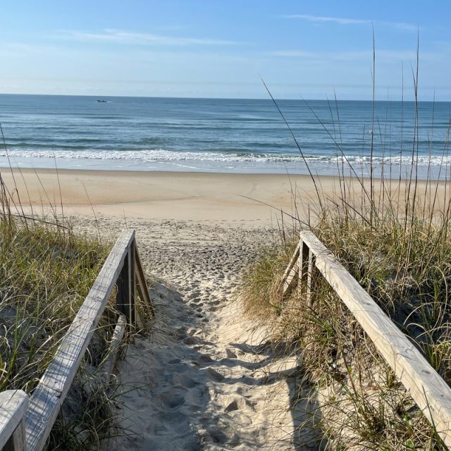 Wooden boardwalk with railings leads through grassy dunes to a sandy beach and calm ocean under a clear sky.