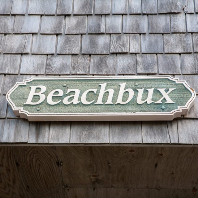 A green sign with the word “Beachbux” mounted on a wooden shingle wall.