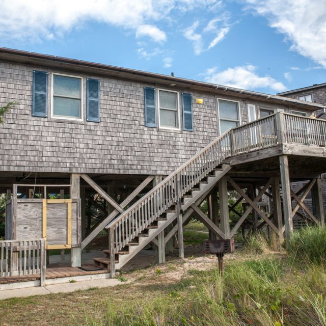 Elevated wooden beach house with shingled siding, large deck, stairs, and grassy area beneath a partly cloudy sky.