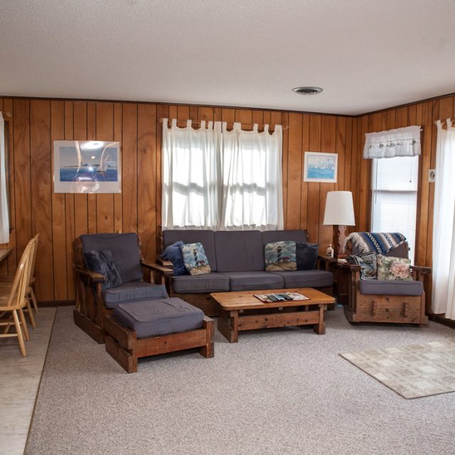 Wood-paneled living room with a couch, armchairs, coffee table, dining area, and door leading to a deck with outdoor view.