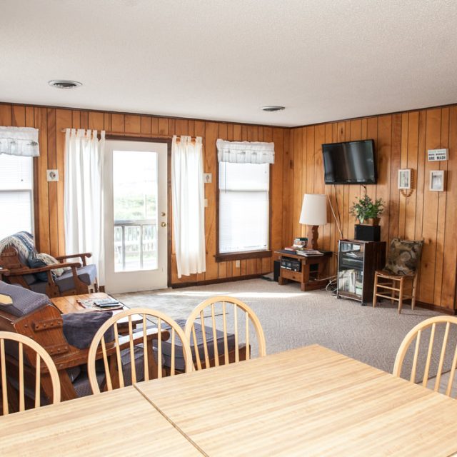 Bright living and dining area with wood-paneled walls, simple furniture, and windows letting in natural light.
